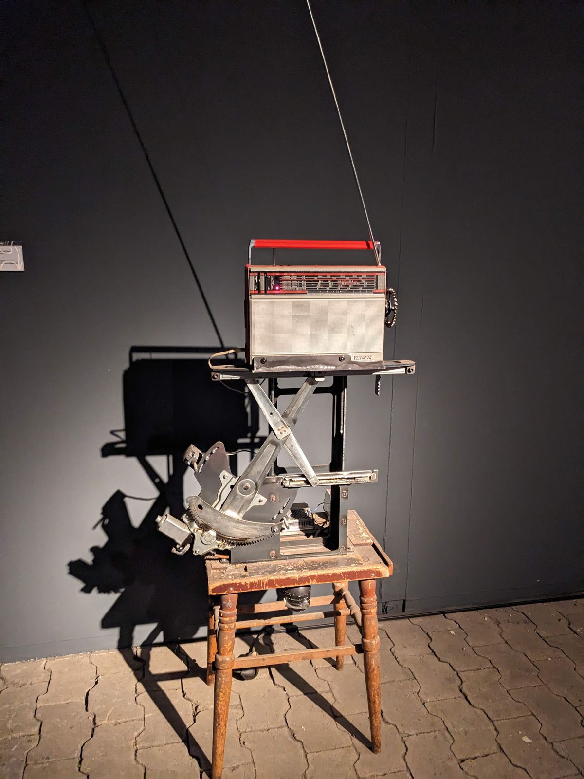 Ein altes rot-weißes Radio mit ausgezogener Antenne steht auf einer mechanischen Scherenhub-Konstruktion aus Metall auf einem alten Holzhocker, an einer schwarzen Wand