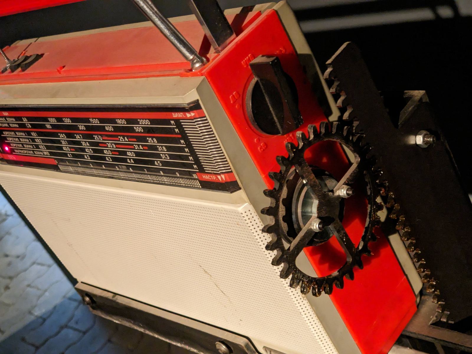 Nahaufnahme des roten Radios mit sichtbarer Senderskala, daneben ein rostiges Zahnrad und eine Fahrradkette als Teil der mechanischen Konstruktion
