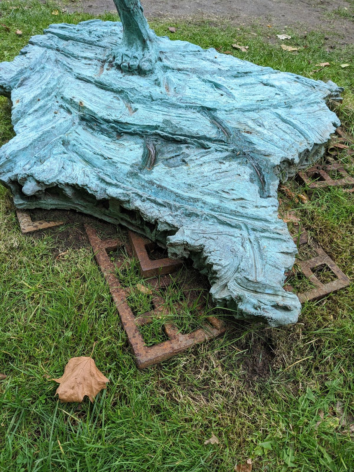 Nahaufnahme des gezackten Bronzesockels der Skulptur mit der eingeprägten Signatur des Künstlers, auf Gras liegend neben einem rostigen Metallgitter