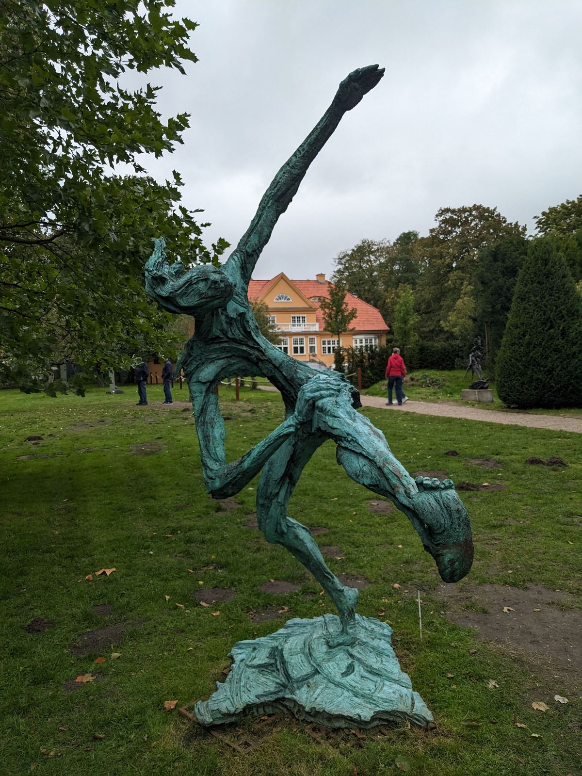 Die Bronzeskulptur „Street Dancer“ von einer anderen Seite gesehen, im Hintergrund ein gelbes Herrenhaus mit rotem Ziegeldach und Besucher im Park