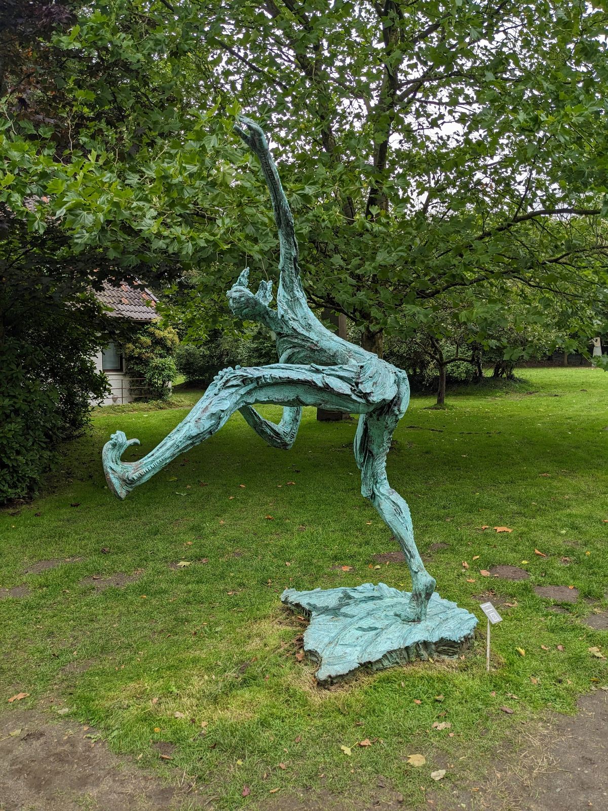 Bronzeskulptur „Street Dancer“ von Richard Brixel im Park der NordArt: eine abstrahierte, tänzerisch bewegte Figur mit weit ausgestreckten Gliedmaßen auf einer gezackten Bronzeplatte, im Hintergrund Bäume und ein kleines Haus