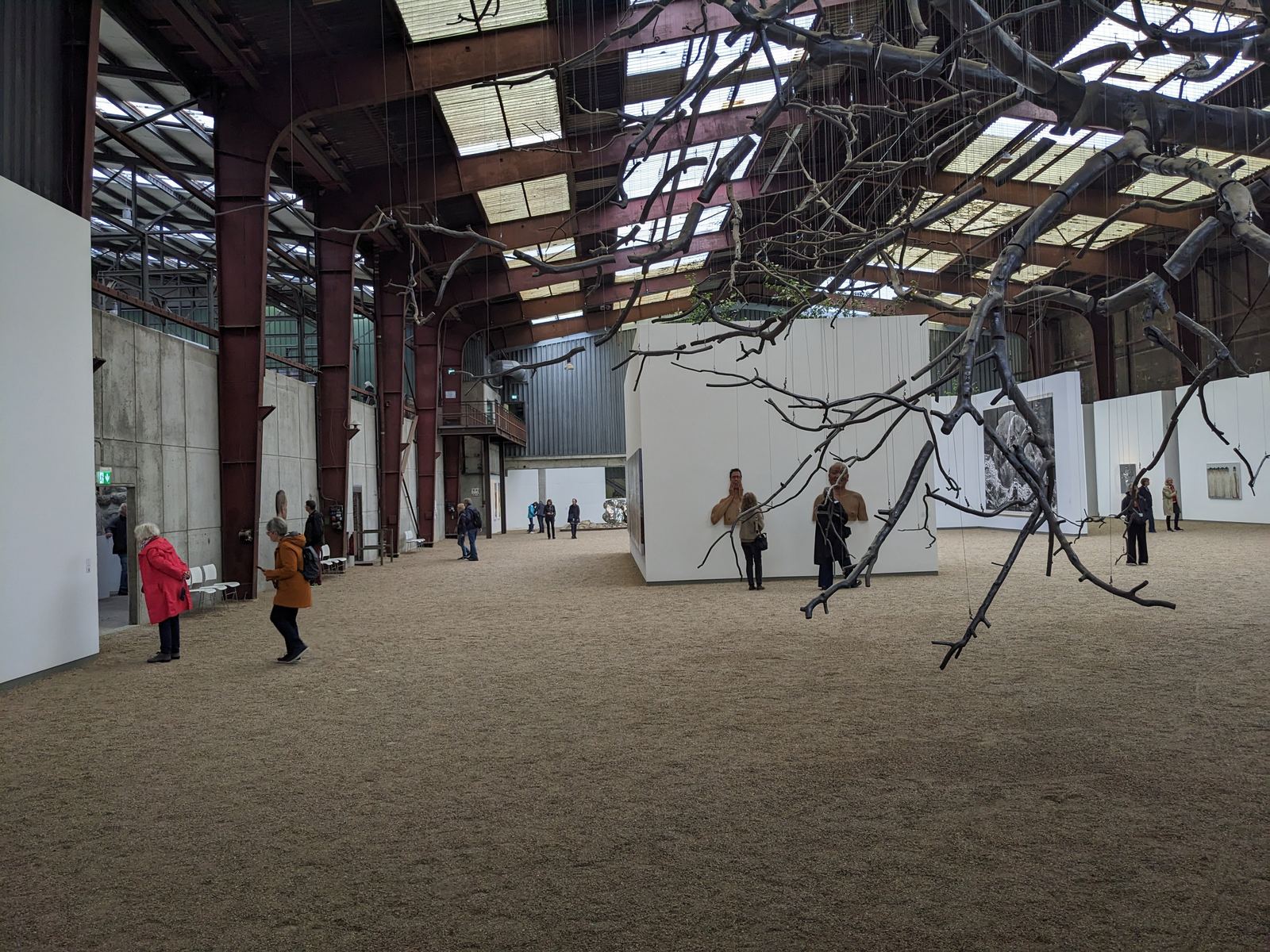 NordArt Carlshütte - Weit verzweigtes, dunkles Astwerk hängt von der Decke einer Industriehalle, darunter Besucher vor einer weißen Wand mit reliefartigen Figuren