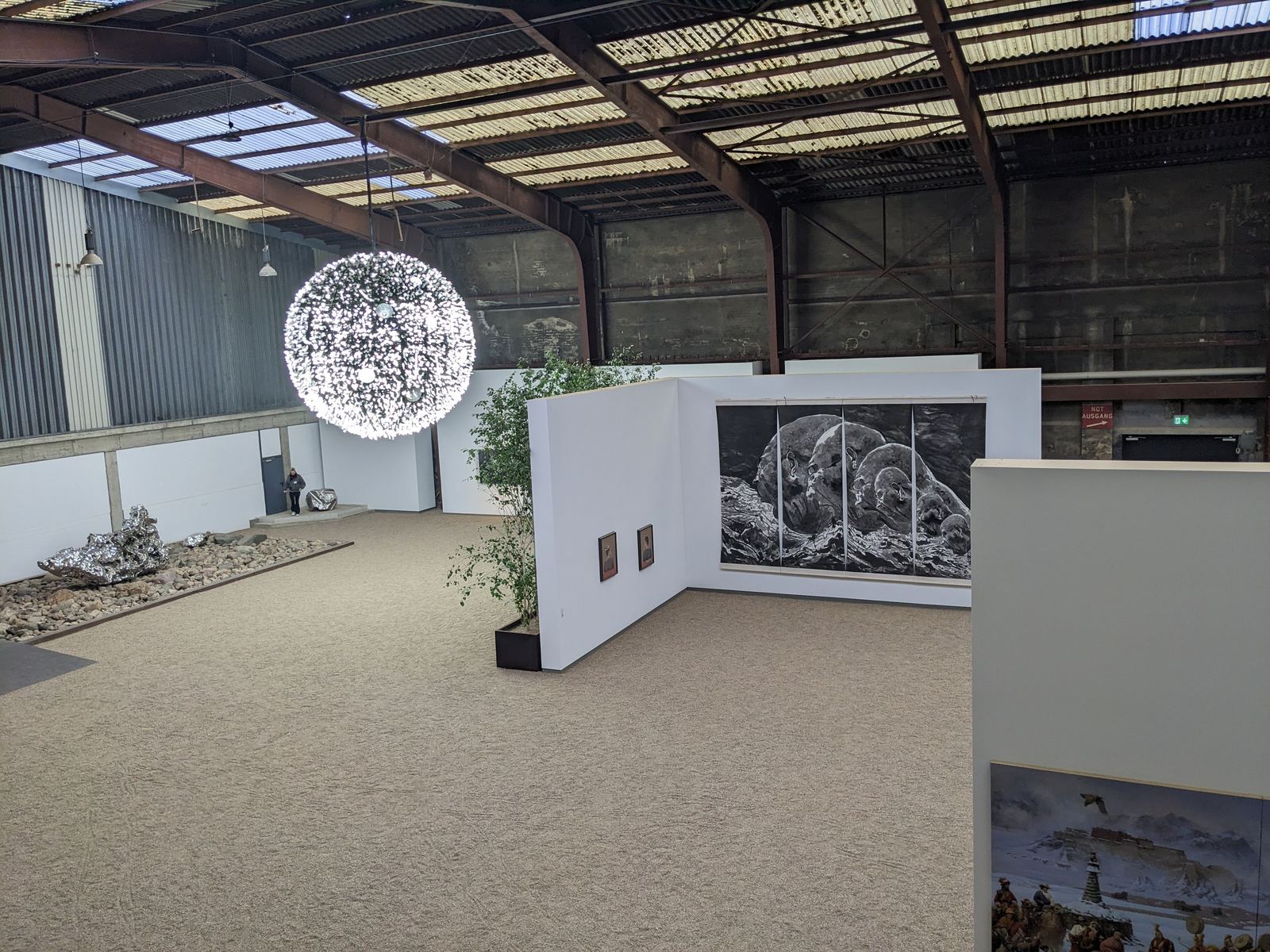 Eine leuchtende Kugel aus unzähligen kleinen Lichtpunkten hängt von der Decke der Halle, daneben Gemälde und ein Kies-Steinhaufen mit einer silbrig glänzenden Skulptur