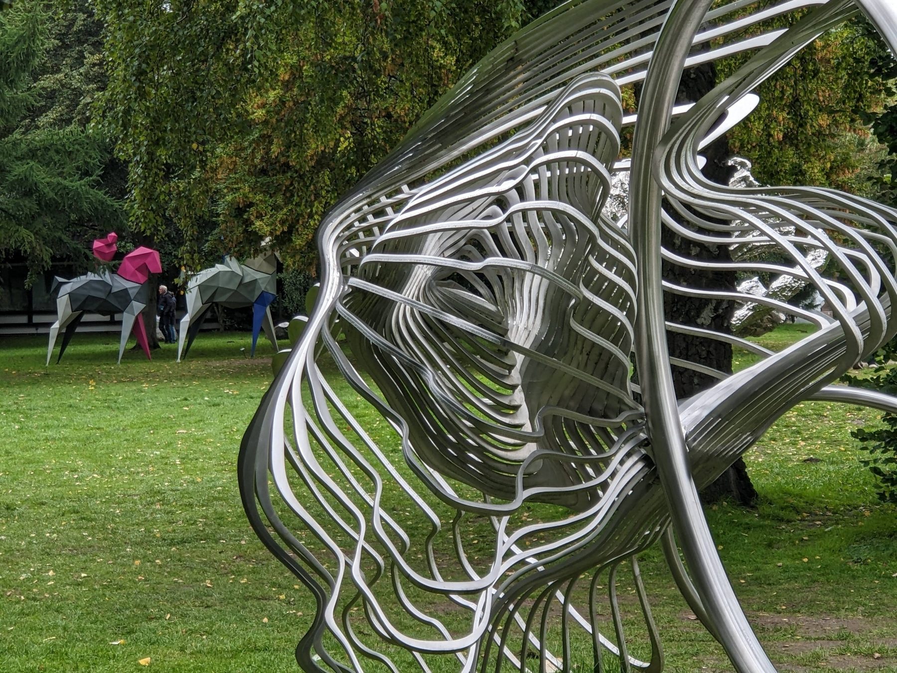 Detailansicht der Edelstahlskulptur Shell von Jan Dostál mit geschwungenen Metalllinien vor Bäumen.