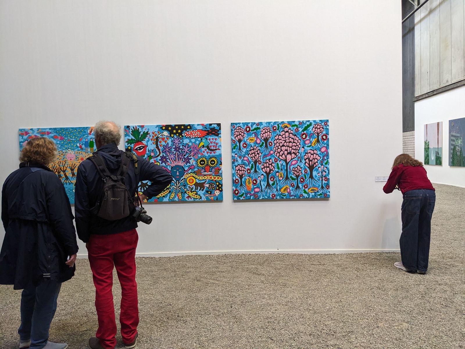 Zwei weitere Gemälde von Marko Mäetamm in Blautönen mit comicartigen Bäumen, Blüten und einem großen blauen Gesicht, Besucher betrachten die Werke in der Ausstellungshalle