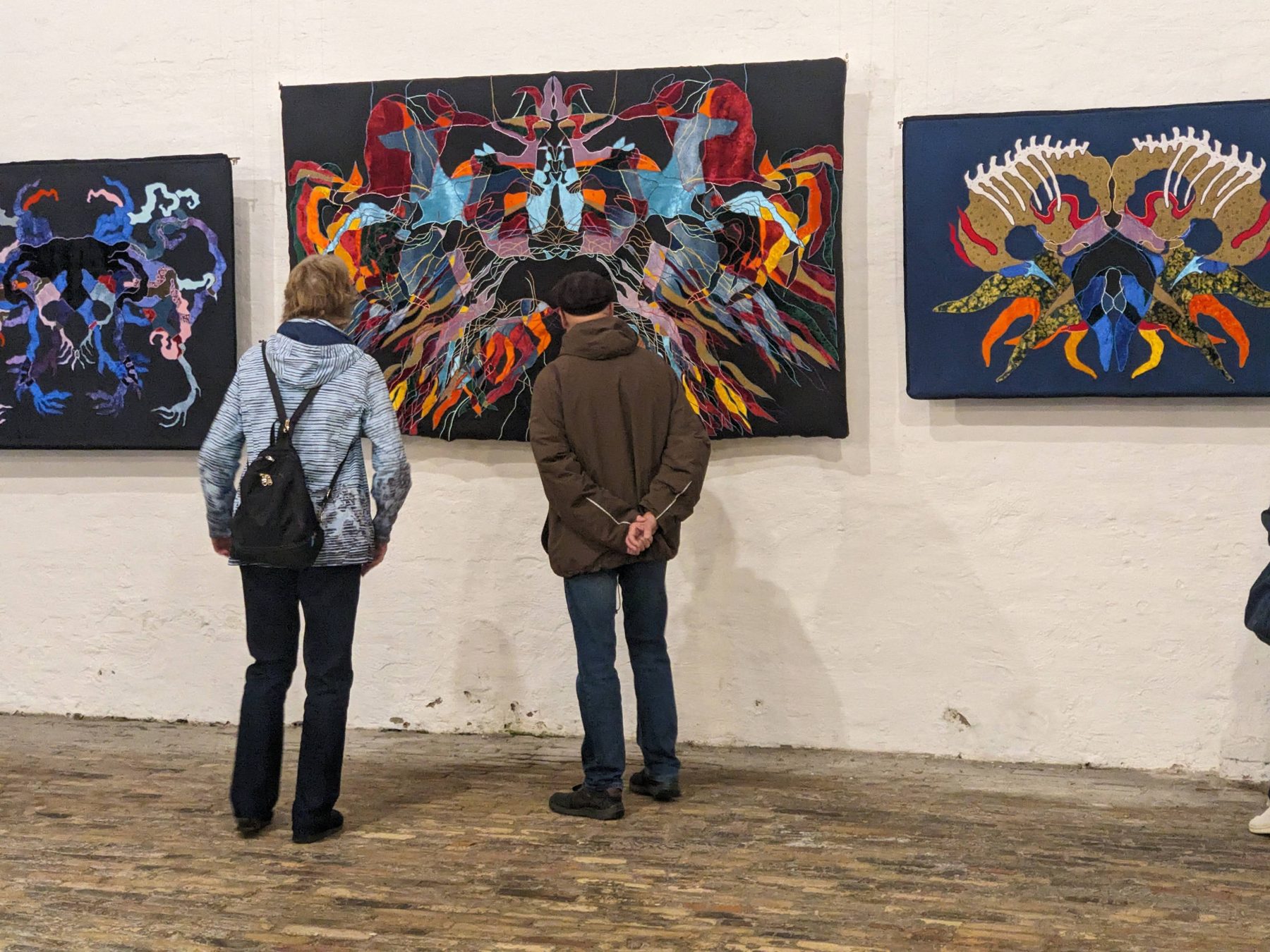 Besucher vor drei farbigen Stickereien von Munisa Kholkhujaeva an einer weißen Ausstellungswand.