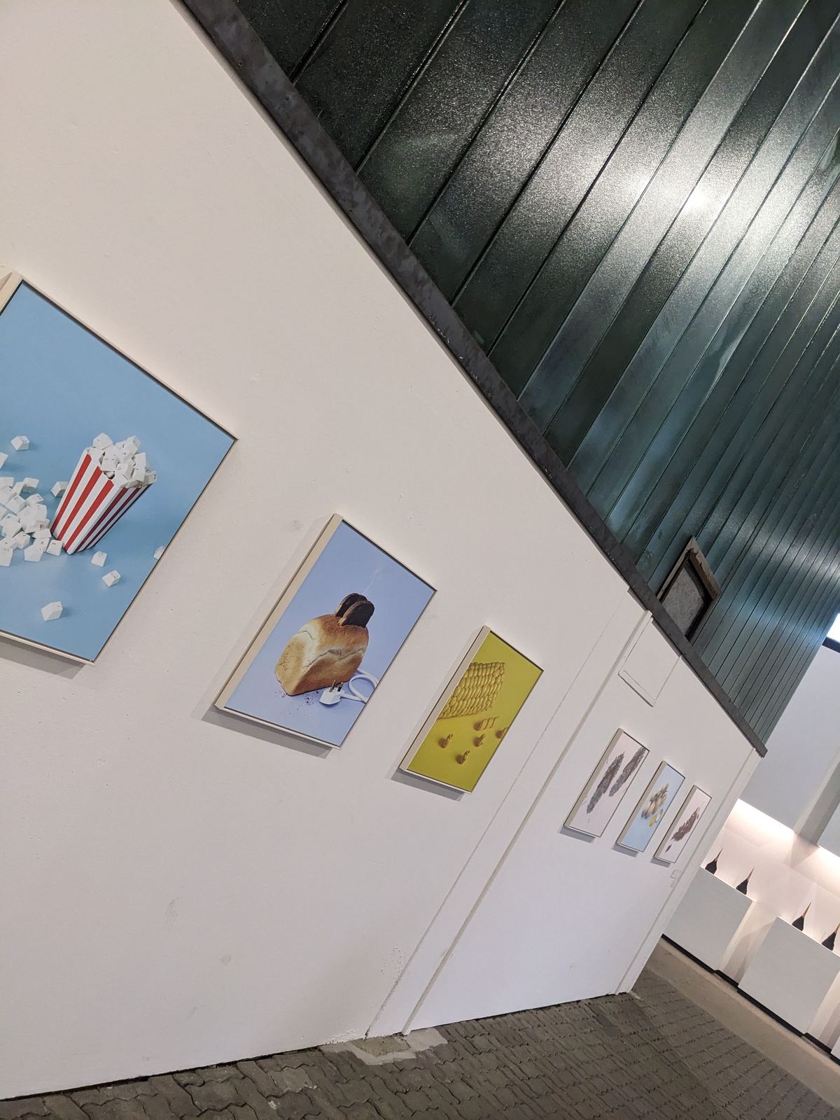 Mehrere Werke der Serie hängen nebeneinander an einer weißen Wand: Popcorn-Tüte, Toastbrot-Toaster und Maiskolben