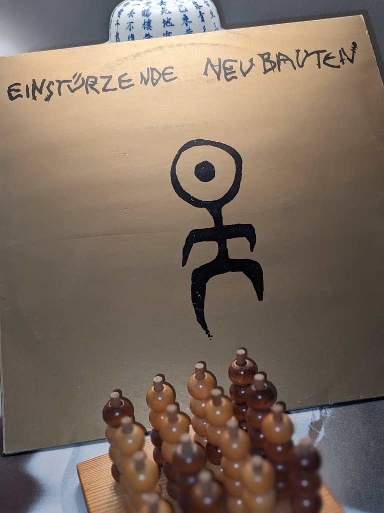 Vorderseite des Plattencovers des Albums Kollaps von Einstürzende Neubauten in Gold mit schwarzem Symbol und Bandaufschrift.