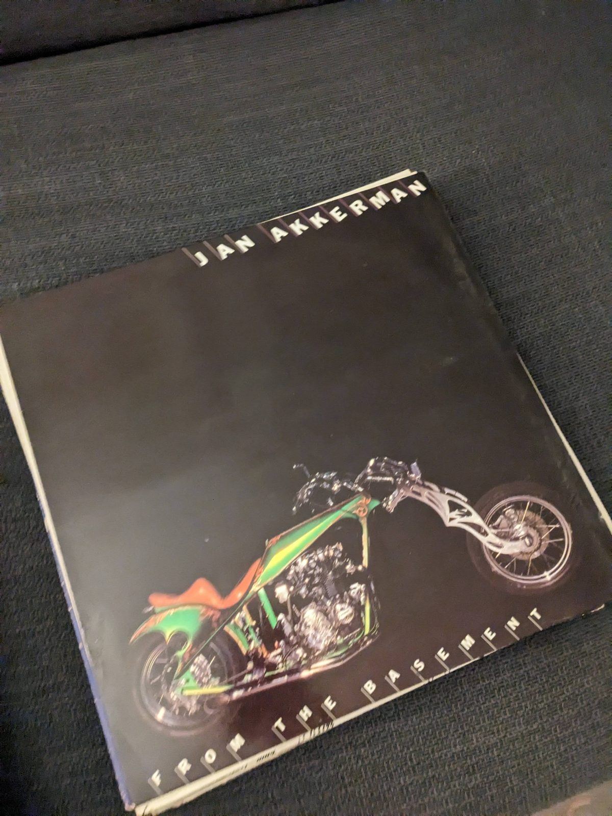 Vorderseite eines Schallplattencovers mit schwarzem Hintergrund, oben beschriftet mit 'JAN AKKERMAN' und unten mit 'FROM THE BASEMENT'. Dazwischen ist ein langgestrecktes Chopper-Motorrad mit Chromteilen und grün-oranger Verkleidung abgebildet.
