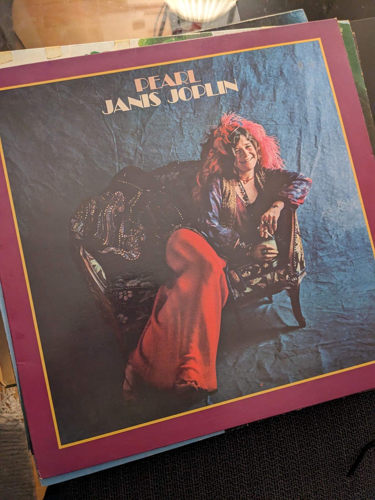 Schallplattencover des Albums Pearl: Janis Joplin sitzt lächelnd auf einem gepolsterten Sessel vor dunklem Hintergrund, trägt eine rote Federboa im Haar, bunte Kleidung und hält ein Glas in der Hand. Das Bild ist von einem breiten purpurroten Rahmen mit goldgelber Konturlinie umgeben. Schallplatten Blog Empfehlung