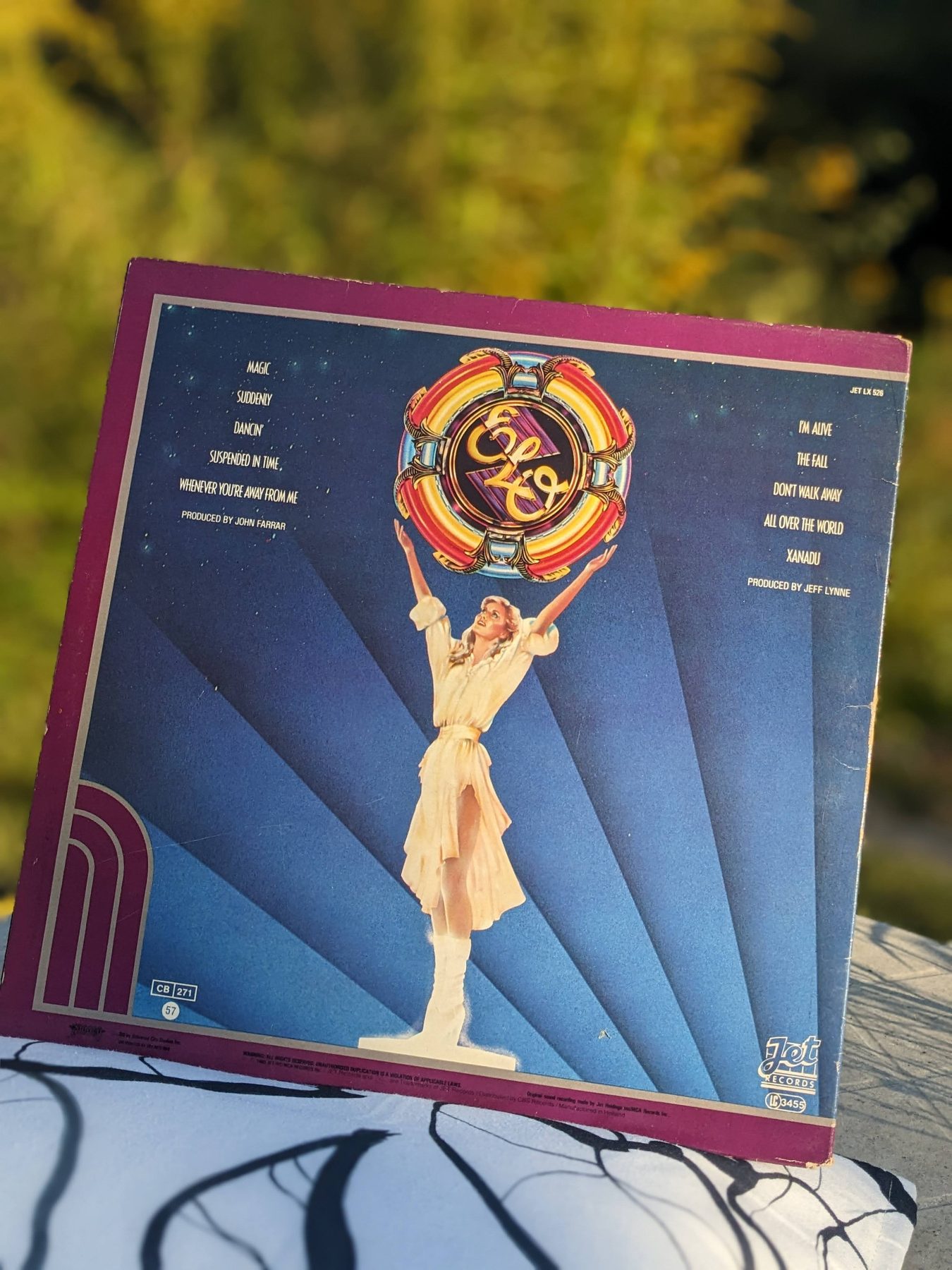 Rückseite der Xanadu-LP im Freien mit blauem Strahlenmotiv, einer ganzfigurigen Darstellung von Olivia Newton-John und dem farbigen ELO-Emblem.